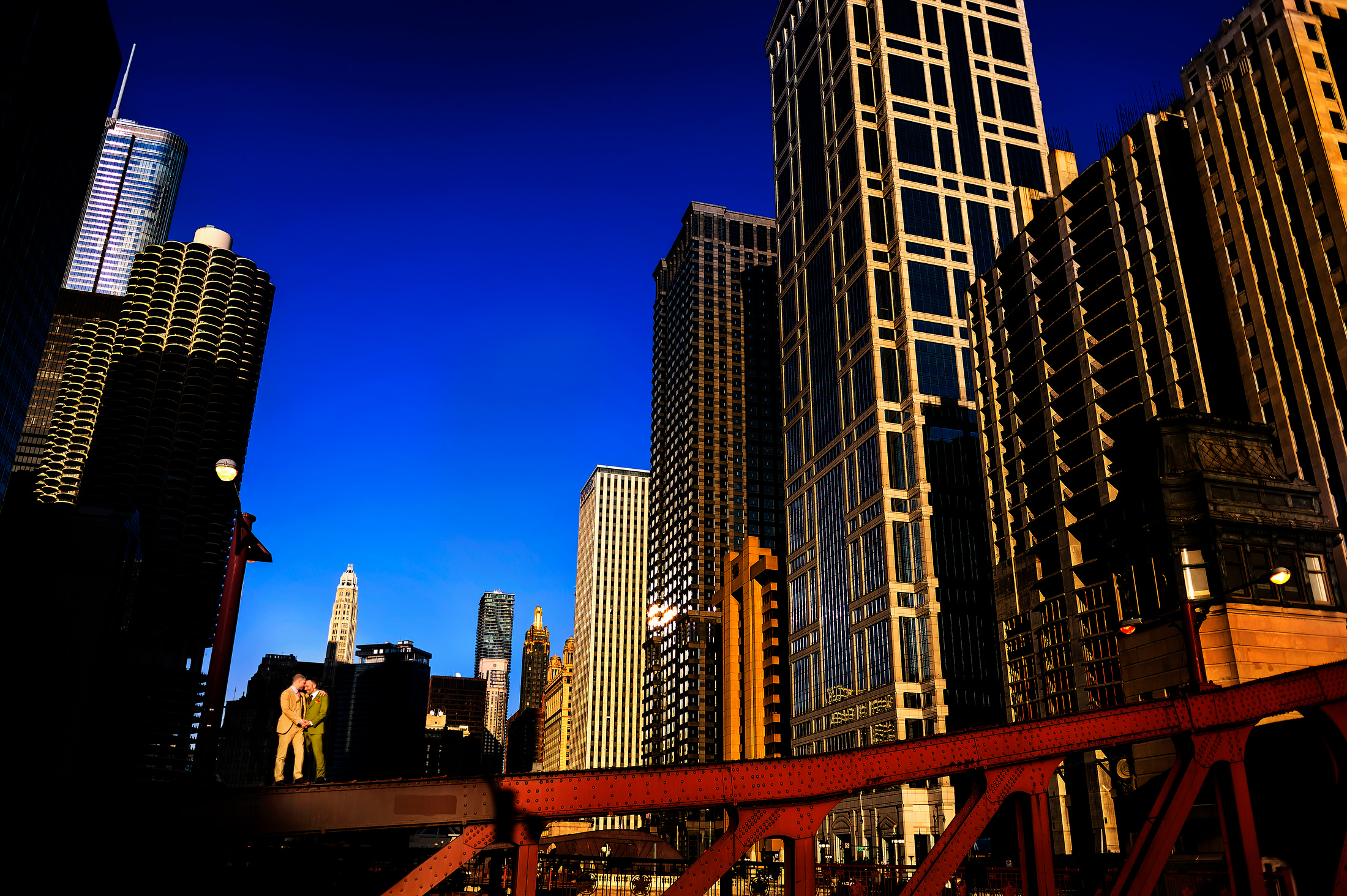 Two grooms stand on bridge in downtown Chicago.